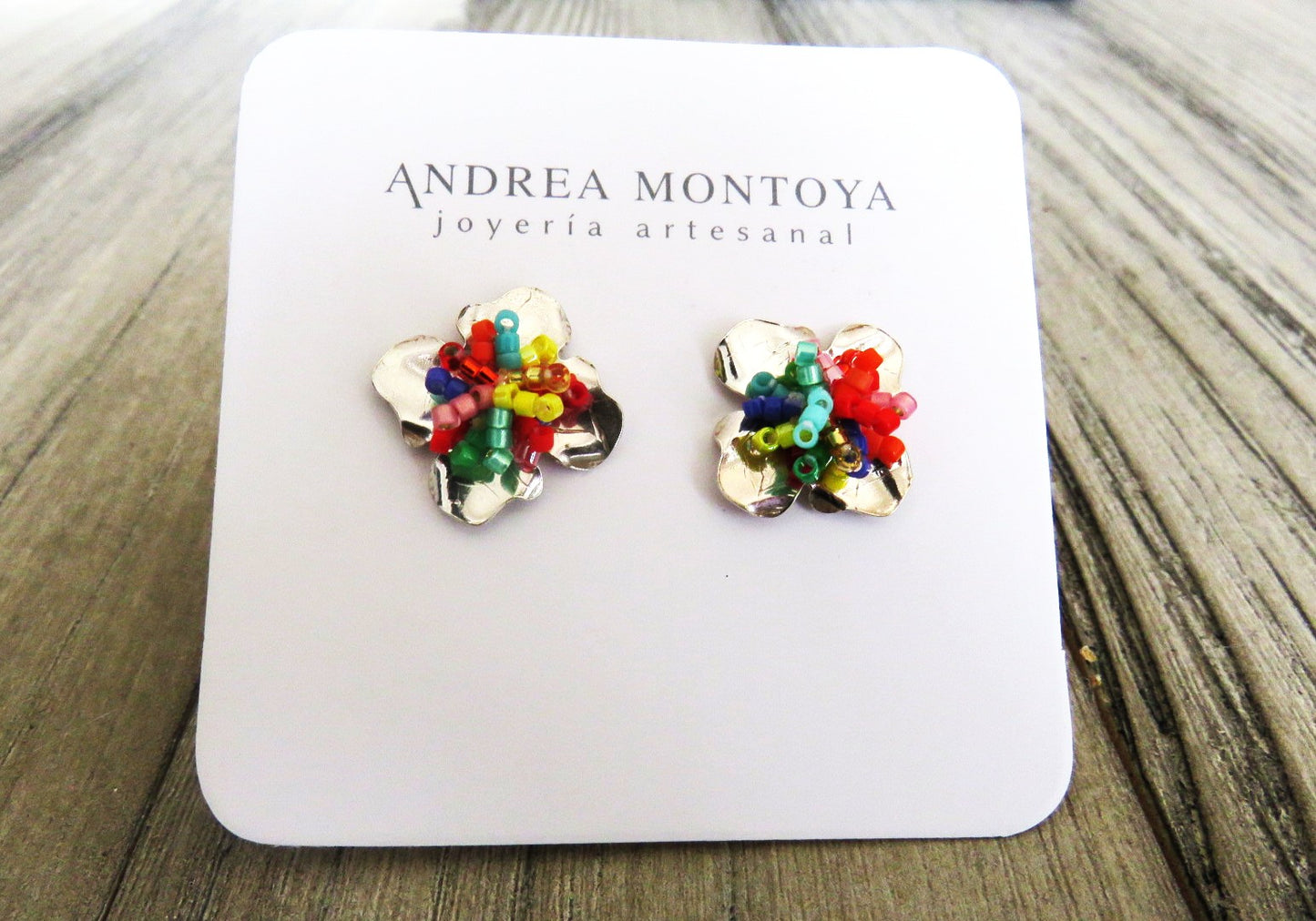 Colorful flower-shaped earrings on a branded card with 'Andrea Montoya' on a wooden surface.