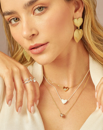 A person wearing a set of gold-colored necklaces with heart-shaped charms and a ring-shaped charm, along with matching earrings and rings.