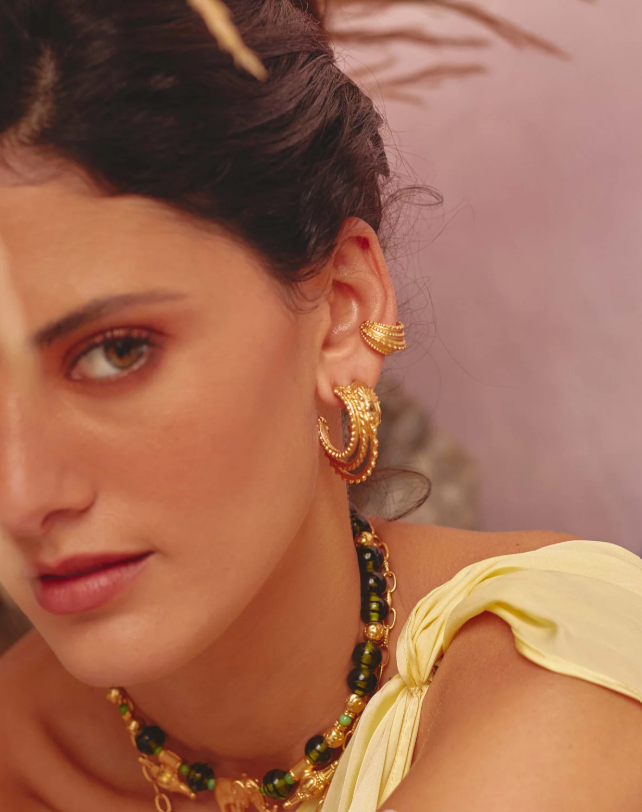 A person wearing a yellow top and gold-colored hoop earrings and an earcuff with intricate designs.