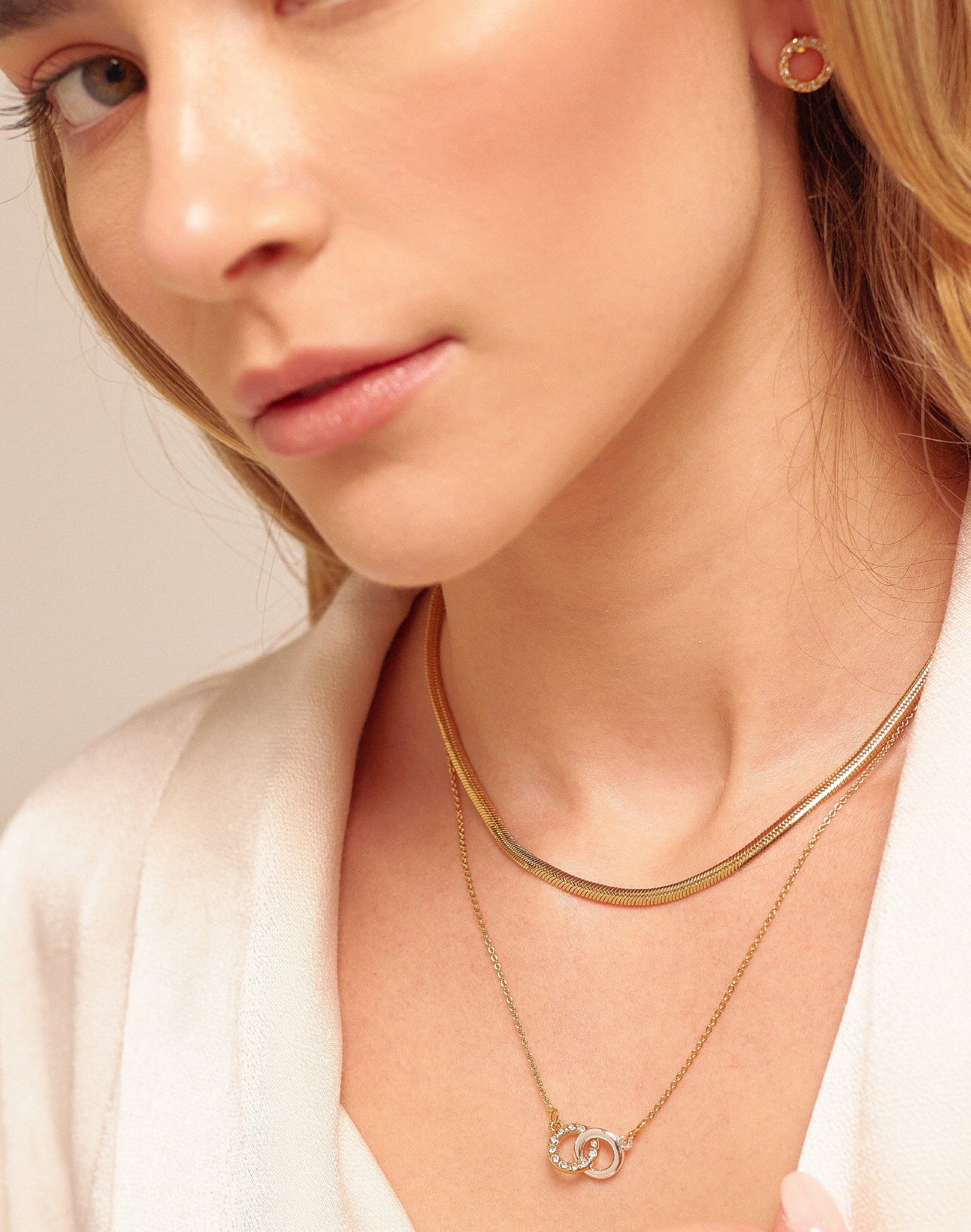 A woman wearing a white shirt, featuring two necklaces with infinity-shaped charms and a pair of hoop earrings.