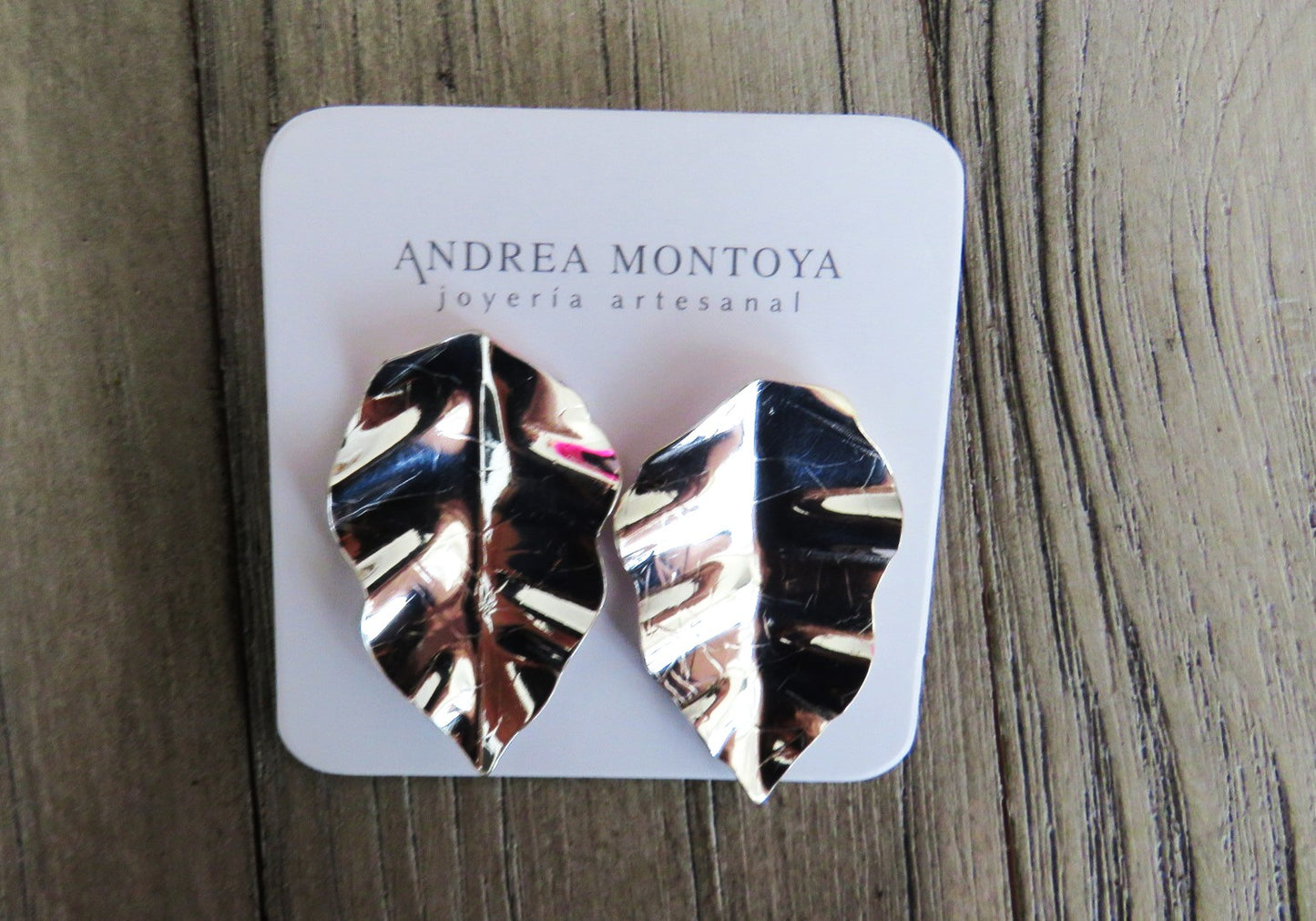 Silver leaf-shaped earrings on a branded card with 'Andrea Montoya' on a wooden surface.