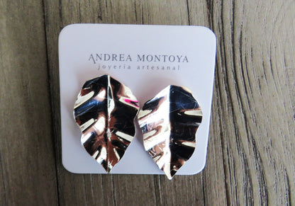 Silver leaf-shaped earrings on a branded card with 'Andrea Montoya' on a wooden surface.