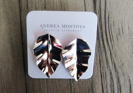 Silver leaf-shaped earrings on a branded card with 'Andrea Montoya' on a wooden surface.