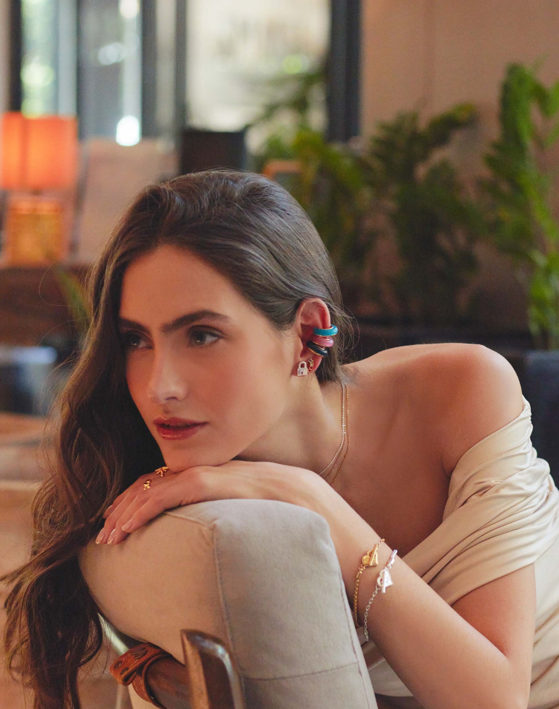 A person wearing a gold-colored ear cuff on one ear and a bracelet on one wrist, with a plant in the background.