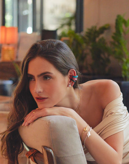 A person wearing a gold-colored ear cuff on one ear and a bracelet on one wrist, with a plant in the background.