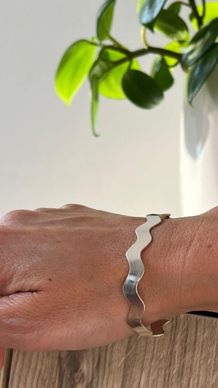 Silver wave-patterned bracelet on a wrist with a blurred plant and light background